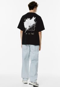 Sort bomulds T-shirt med et hvidt splash-graphic på ryggen. Modellen har parret den med lyseblå baggy jeans og sneakers.