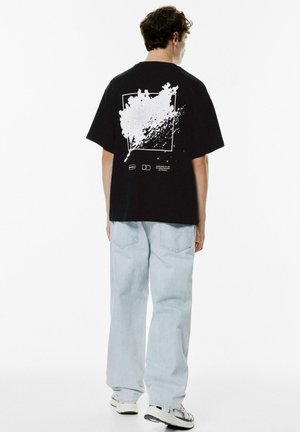 Sort bomulds T-shirt med et hvidt splash-graphic på ryggen. Modellen har parret den med lyseblå baggy jeans og sneakers.