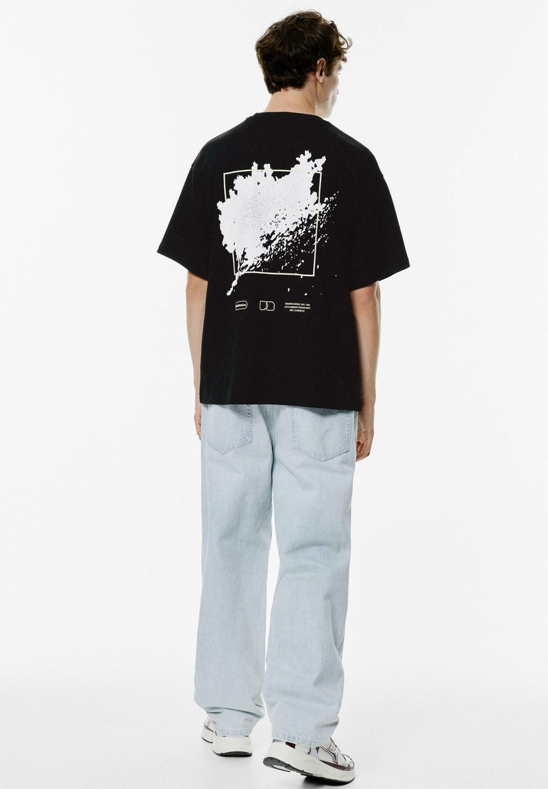 Sort bomulds T-shirt med et hvidt splash-graphic på ryggen. Modellen har parret den med lyseblå baggy jeans og sneakers.