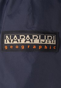 Il tessuto in nylon blu navy presenta una piccola toppa nera rettangolare con il testo in rilievo "NAPAPIJRI" in bianco e "geographic" in arancione sotto.