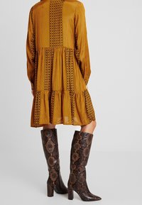 Langes gelbes Kleid mit vertikalen Streifen und floralen Mustern, kombiniert mit knielangen braunen Schlangenlederstiefeln mit Blockabsatz.