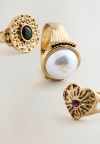 Drei Goldringe: einer mit einer großen weißen Perle, ein anderer mit einem schwarzen Stein und kunstvollem Design, sowie ein herzförmiger Ring mit rotem Akzent.