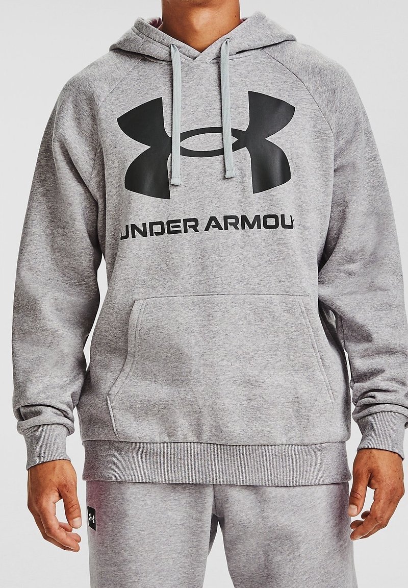 Homme portant un sweat à capuche gris Under Armour et un pantalon de survêtement gris assorti, mains détendues le long du corps, visible du cou jusqu’aux hanches.