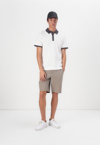 Λευκό πόλο μπλουζάκι με σκούρο γιακά, μπεζ σορτς και λευκά αθλητικά παπούτσια. Το πόλο έχει μία διακριτική υφή και ένα λογότυπο στο στήθος.