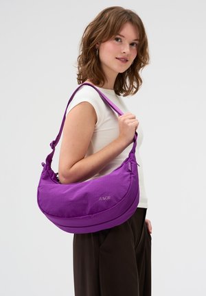 Junge Frau trägt ein weißes Oberteil und dunkle Hose, hält eine große lila Umhängetasche und steht vor einem einfarbigen hellen Hintergrund.