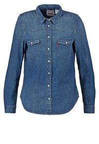 Chemise en denim bleu à manches longues avec un col pointu, deux poches poitrine et des boutons argentés. Texture douce avec une coupe classique et un ourlet arrondi.