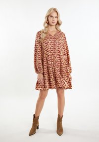 Rochie cu un model roșu și auriu, cu decolteu în V și mâneci lungi, având o fustă evazată și țesătură texturată. Pur și simplu cu bocanci maro până la gleznă.