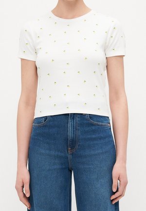 T-shirt blanc court avec un petit motif de fruits jaunes, porté avec un jean bleu taille haute, visible des épaules aux hanches.