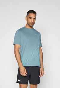Camiseta atlética de color azul claro con mangas cortas, que presenta rayas horizontales. El material es suave y tiene un corte relajado. Pantalones cortos negros con logo.