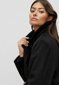 Svart ullblandad kappa med hög krage, med en slät textur och minimalistisk design, delvis som täcker modellens hand.