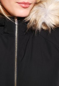 Černá bunda s vysokým límcem, vybavená lesklým zlatým zipem a odnímatelným umělým kožíškem kolem kapuce.