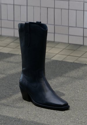 Korkea musta nahkainen cowboy-saapas, jossa on terävä kärki ja vinosti asetettu keskipitkä korko, seisoo päällystettyä seinää vasten jalkakäytävällä.