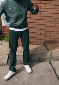 Person iført mørkegrønne bukser, hvide sneakers og en mørkegrøn zip-up jakke, som står på et betonfortov ved en mur af mursten.