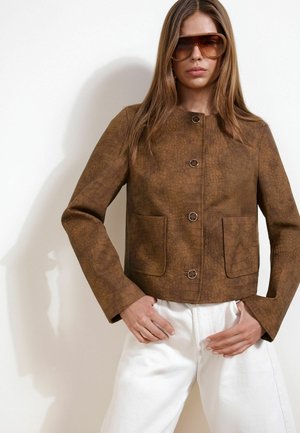 Femme portant une veste marron texturée avec des poches avant, un pantalon blanc et des lunettes de soleil marron surdimensionnées, debout les mains près des hanches.