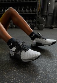 Osoba v černobílých sportovních botách a černých ponožkách sedící na podlaze v tělocvičně s jednoručními činkami v pozadí.