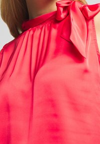 Blouse en satin rouge avec un col montant, présentant un détail froncé et un nœud décoratif à l'épaule. Texture lisse et coupe ajustée.