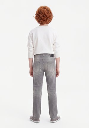 Enfant aux cheveux roux bouclés, debout de dos, portant une chemise blanche à manches longues, un jean gris avec des éclaboussures de peinture et des baskets grises.