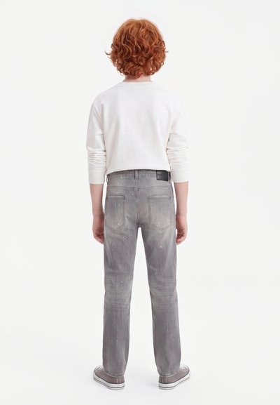 Enfant aux cheveux roux bouclés, debout de dos, portant une chemise blanche à manches longues, un jean gris avec des éclaboussures de peinture et des baskets grises.