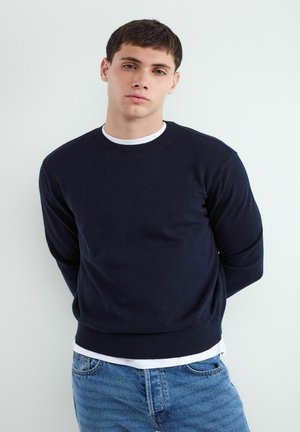 Junger Mann steht an einer weißen Wand, trägt einen dunkelblauen Pullover über einem weißen Hemd und blaue Jeans, Hände hinter dem Rücken, neutraler Gesichtsausdruck.