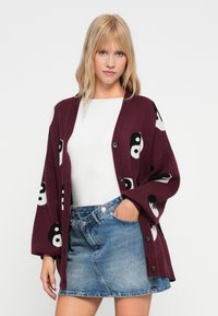 Burgundy színű kardigán, yin-yang mintákkal, puha anyagból készült, laza fazonú, hosszú ujjú, gombos záródással. Fehér felső felett viselik.