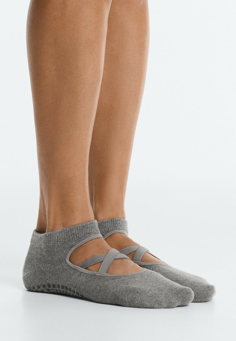 Pieds portant des chaussettes grises courtes avec des bandes élastiques croisées et des points antidérapants sous la semelle, posés sur une surface blanche lisse.