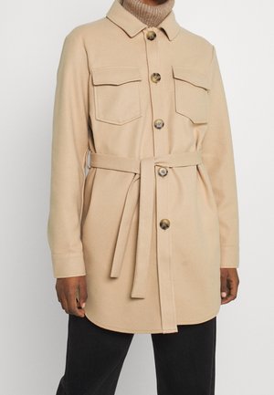 Person iført beige bæltejakke med knaplukning og brystlommer med klap over en brun rullekrave og sorte bukser.