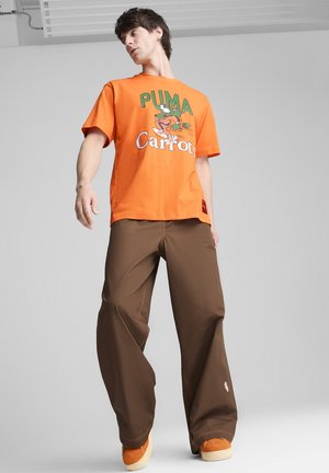 Pomarańczowa bawełniana koszulka z zieloną grafiką "PUMA Carrots"; zestawiona z brązowymi, luźnymi spodniami; pomarańczowe buty z kremowymi akcentami.