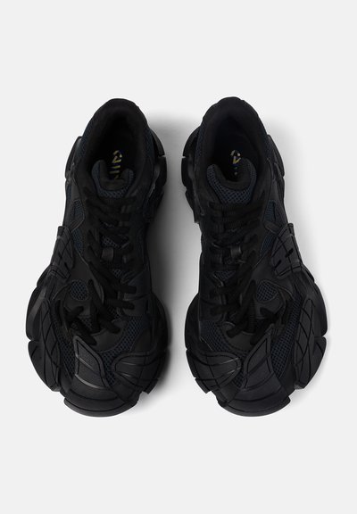 Coppia di sneakers nere con suola spessa e pannelli testurizzati, viste dall'alto su uno sfondo bianco.
