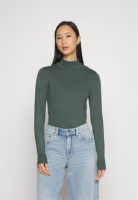 Zeleni pulover s turtleneck ovratnikom, izdelan iz fine pletenine, z dolgimi rokavi in oprijetim krojem, v kombinaciji s svetlo modrimi hlačami z visokim pasom.
