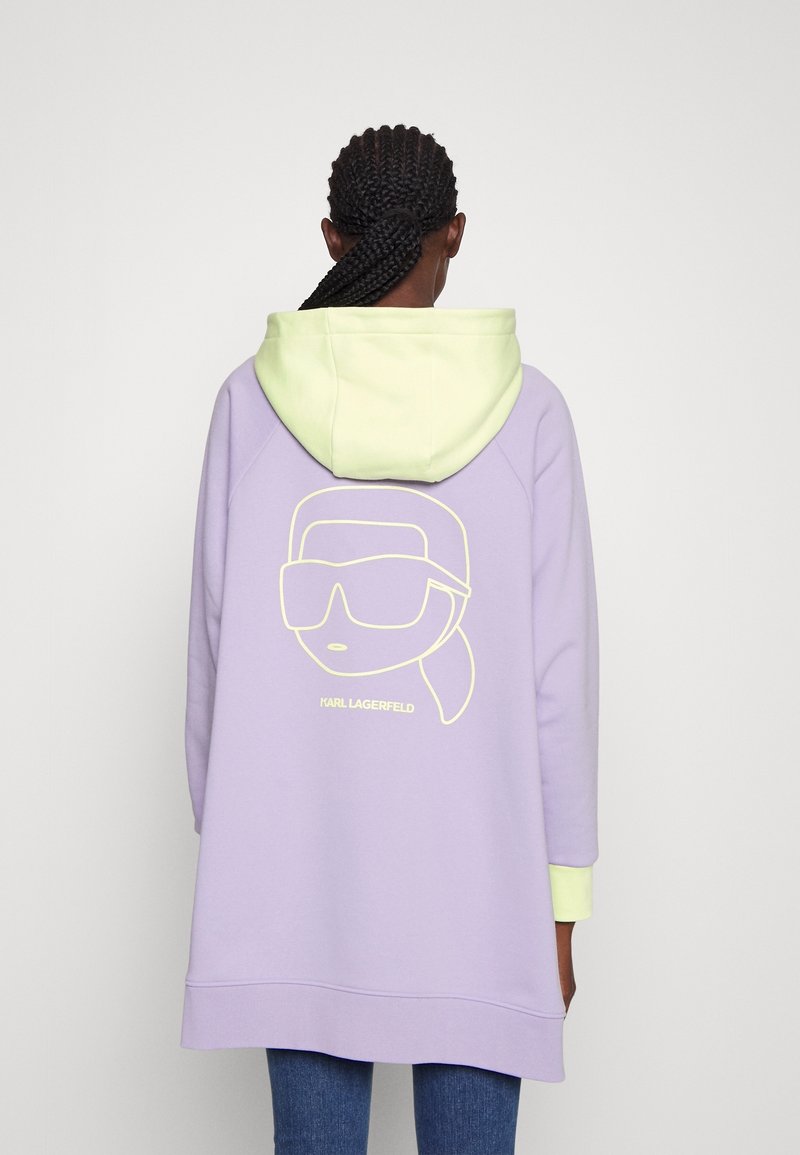 Personne avec des cheveux tressés portant un sweat à capuche lavande avec une capuche jaune clair et un design minimaliste de visage avec des lunettes de soleil dans le dos.