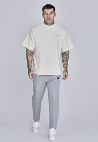 Beige oversize t-shirt med korta ärmar, ljusgrå figurnära byxor och vita sneakers. Har tatueringar på armarna och en liten logotyp på tröjan.
