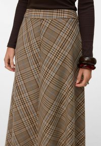 Femme portant une jupe à carreaux marron et un haut à manches longues foncé, avec de larges bracelets marron et une bague argentée à la main droite.