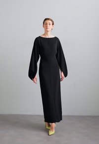 Black long-sleeve dress with a relaxed fit, featuring a boat neckline and flared sleeves. Worn with yellow high-heeled shoes against a neutral background.