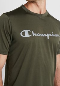 Olivově zelené tričko s krátkým rukávem vyrobené z texturované látky, s velkým bílým logem "Champion" přes hrudník.