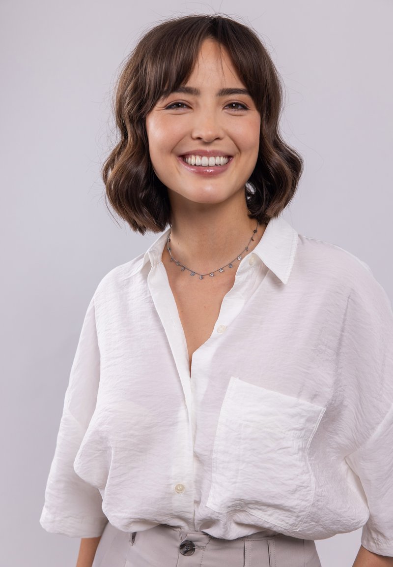 Weißes Hemd mit strukturiertem Finish, ausgestattet mit einer linken Brusttasche und lockeren Ärmeln. Getragen mit hellgrauen, hochgeschnittenen Hosen.