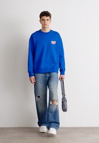 Sweatshirt azul brilhante com gráfico "VERY" no peito, combinado com jeans claros desgastados com rasgos e ténis brancos. A carregar uma mala cinza.