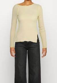 Pitkähiha, vaalea paita, jossa on veneen kaula-aukko ja kontrastivärinen keltainen ommel, hieman epäsymmetrinen helma ja vartalonmyötäinen siluetti.