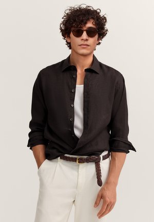 Mann mit lockigem Haar, der dunkle Sonnenbrillen trägt, ein aufgeknöpftes schwarzes Hemd über einem weißen Tanktop, weiße Hose und braunen geflochtenen Gürtel mit den Händen in den Taschen.