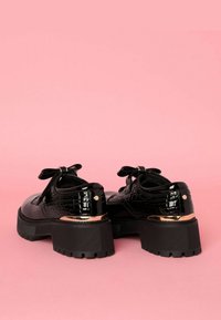Scarpe in pelle nera lucida con texture a coccodrillo, dotate di una suola spessa, dettagli a fiocco e una rifinitura dorata metallica sul tacco.