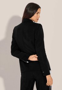 Schwarze Jacke mit taillierter Passform, mit Reverskragen, Ein-Knopf-Verschluss und subtiler Glanzoberfläche; kombiniert mit schwarzen Hosen.