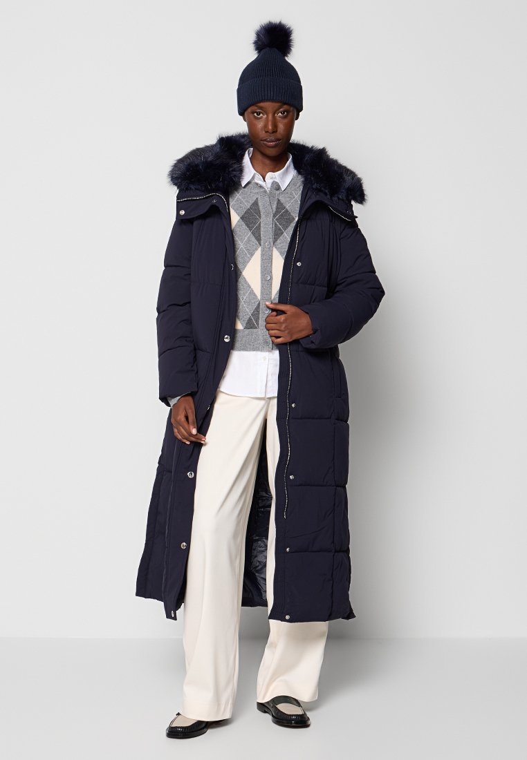 Cappotto lungo imbottito blu navy con cappuccio foderato in pelliccia, dotato di chiusure a scatto, indossato sopra un maglione grigio a rombi e pantaloni color crema.
