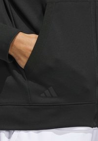 Black athletic hoodie featuring a side pocket and a subtle tonal adidas logo near the hem, made from a smooth, stretchy fabric.