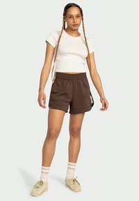 Brune fleece-shorts med elastisk midje, sidelommer og et dekorativt detaljer på siden. Kombinert med en tettsittende hvit t-skjorte.