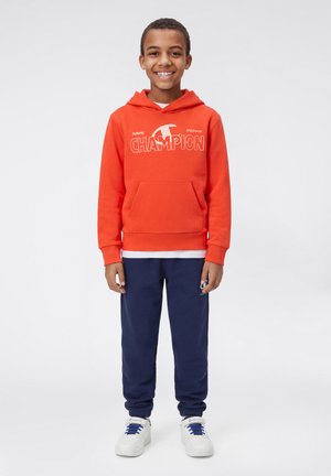 Felpa con cappuccio arancione con logo "CHAMPION" bianco, tasca a marsupio, abbinata a pantaloni della tuta blu navy e sneakers bianche con lacci blu.