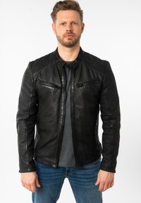 Giacca di pelle nera con tasche con cerniera, dettagli a costine sulle spalle, colletto alto e taglio aderente. Indossata sopra una maglietta grigia e dei jeans blu.
