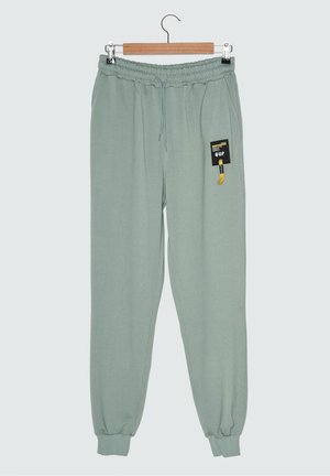 Hellgrüne Jogginghose mit elastischem Bund, Kordelzug und Bündchen, mit einem schwarz-gelben Patch am rechten Oberschenkel, an einem Holzbügel hängend.