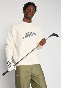 Krémová mikina s logom "Malbon" nosená s olivovými nohavicami. Osoba drží golfovú palicu a má na sebe bielu golfovú rukavicu.