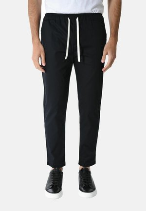 Uomo che indossa pantaloni neri con coulisse e sneakers nere con suole bianche, in piedi davanti a uno sfondo bianco uniforme.
