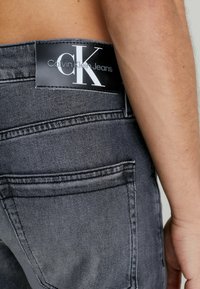 Grå denimjeans med en framträdande svart läderetikett som har "Calvin Klein Jeans"-logotypen, som framhäver sömnads- och fickdetaljer.