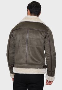 Veste en daim marron avec un col et une doublure en moumoute crème. Comprend des panneaux en cuir verticaux et des sangles ajustables à la taille.